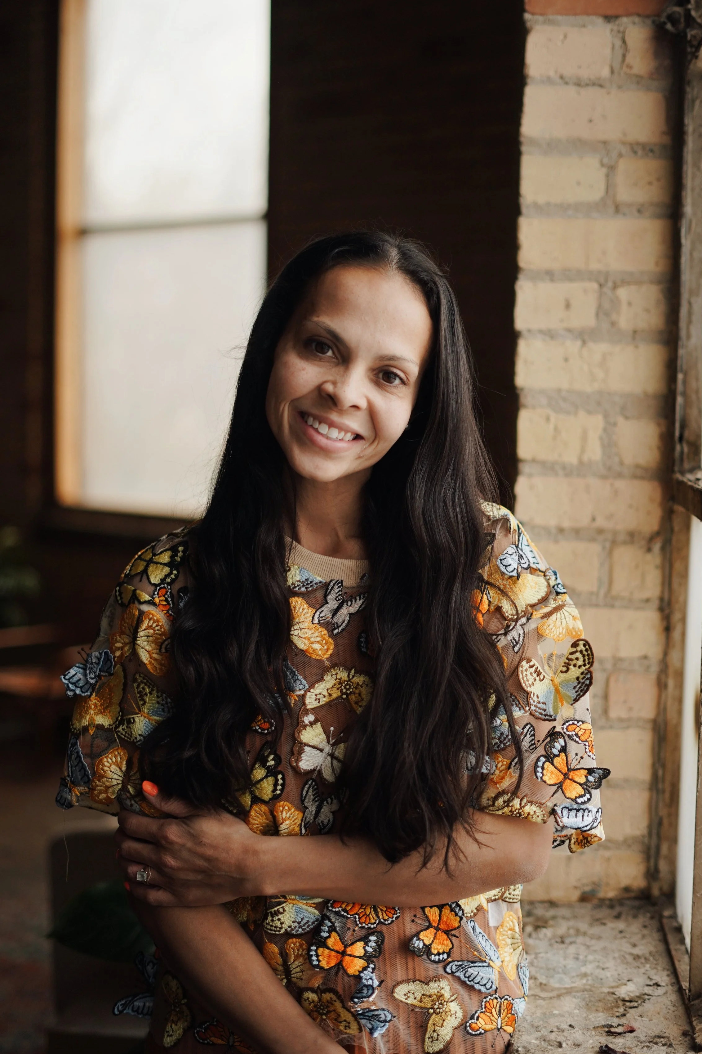 A woman with long dark hair smiling, wearing a sheer fabric top with butterfly embroidery, standing indoors near a window and brick wall.