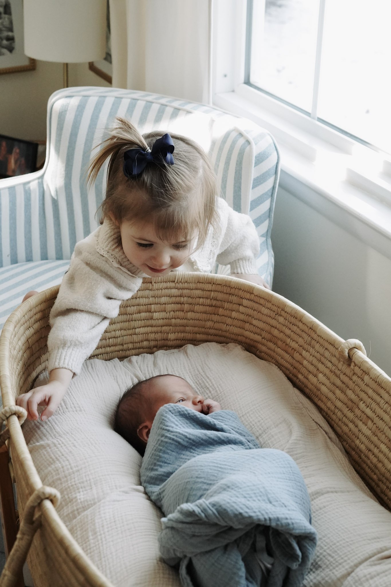 A young girl with a bow in her hair leaning over a bassinet and looking at a newborn baby wrapped in a blanket.