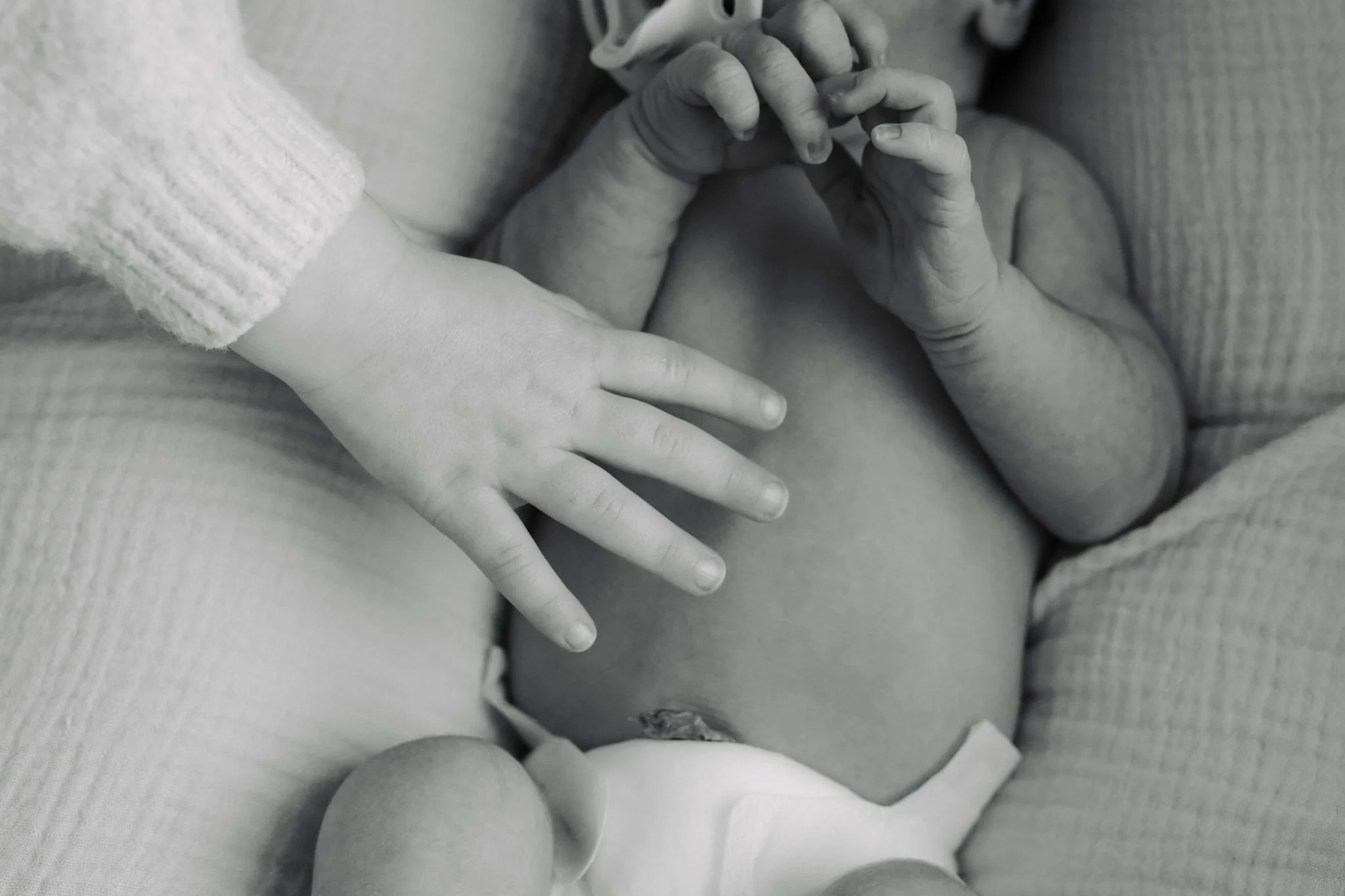Close-up black and white photo of a newborn baby's hand touching an adult's finger, with the baby's body partially visible and the adult wearing a sweater.
