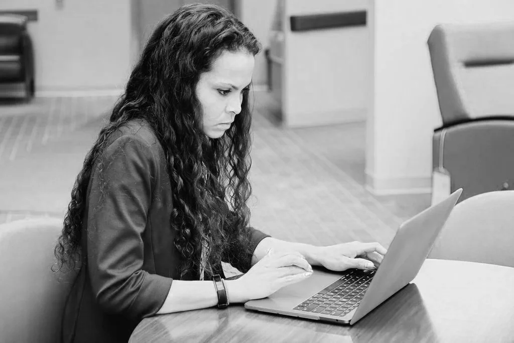 A woman with long curly hair working on a laptop at a table in an office or waiting area.