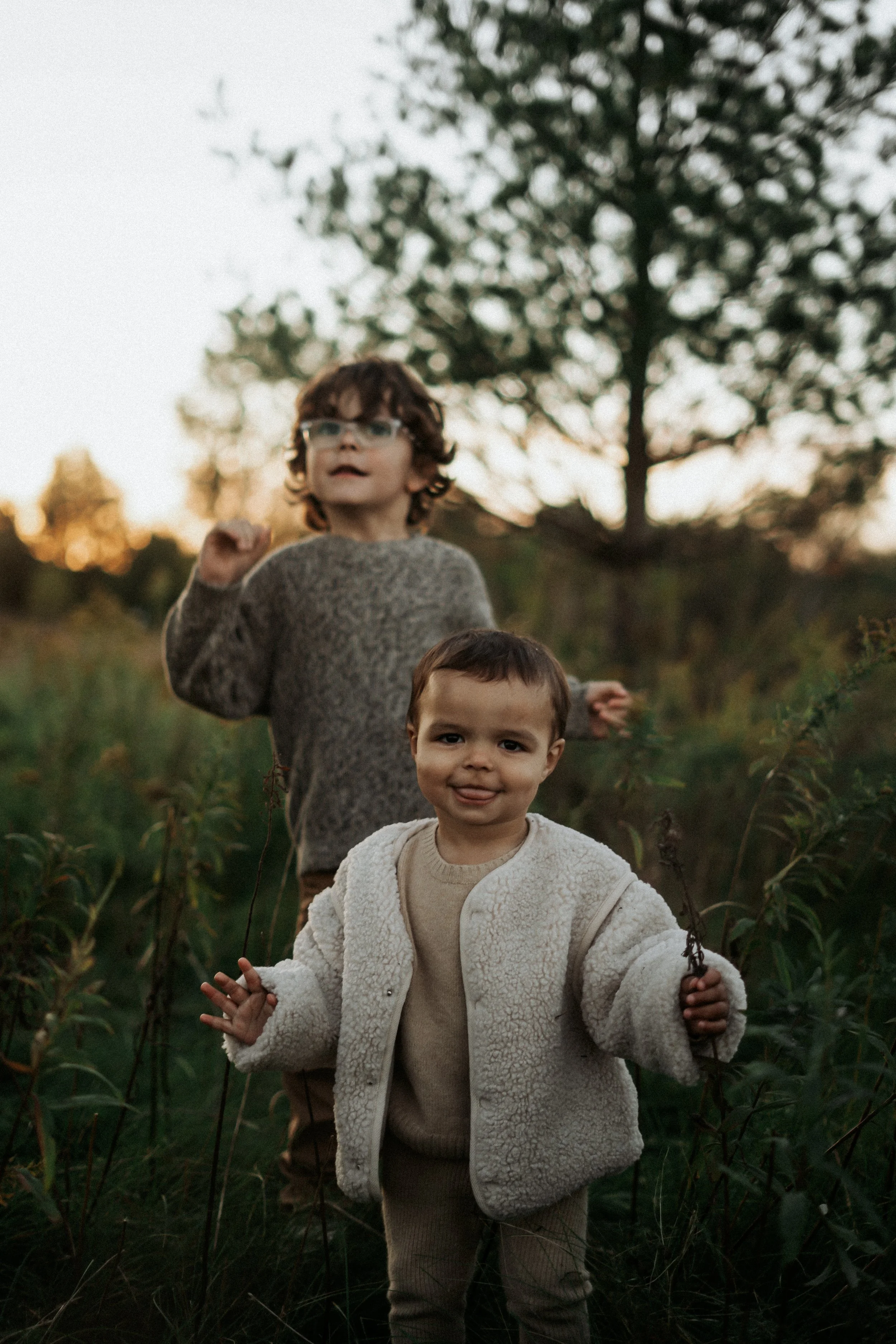 Two young children standing outdoors in a field with tall grass and bushes during sunset, one in the foreground smiling and sticking out tongue, the other in the background raising one hand.