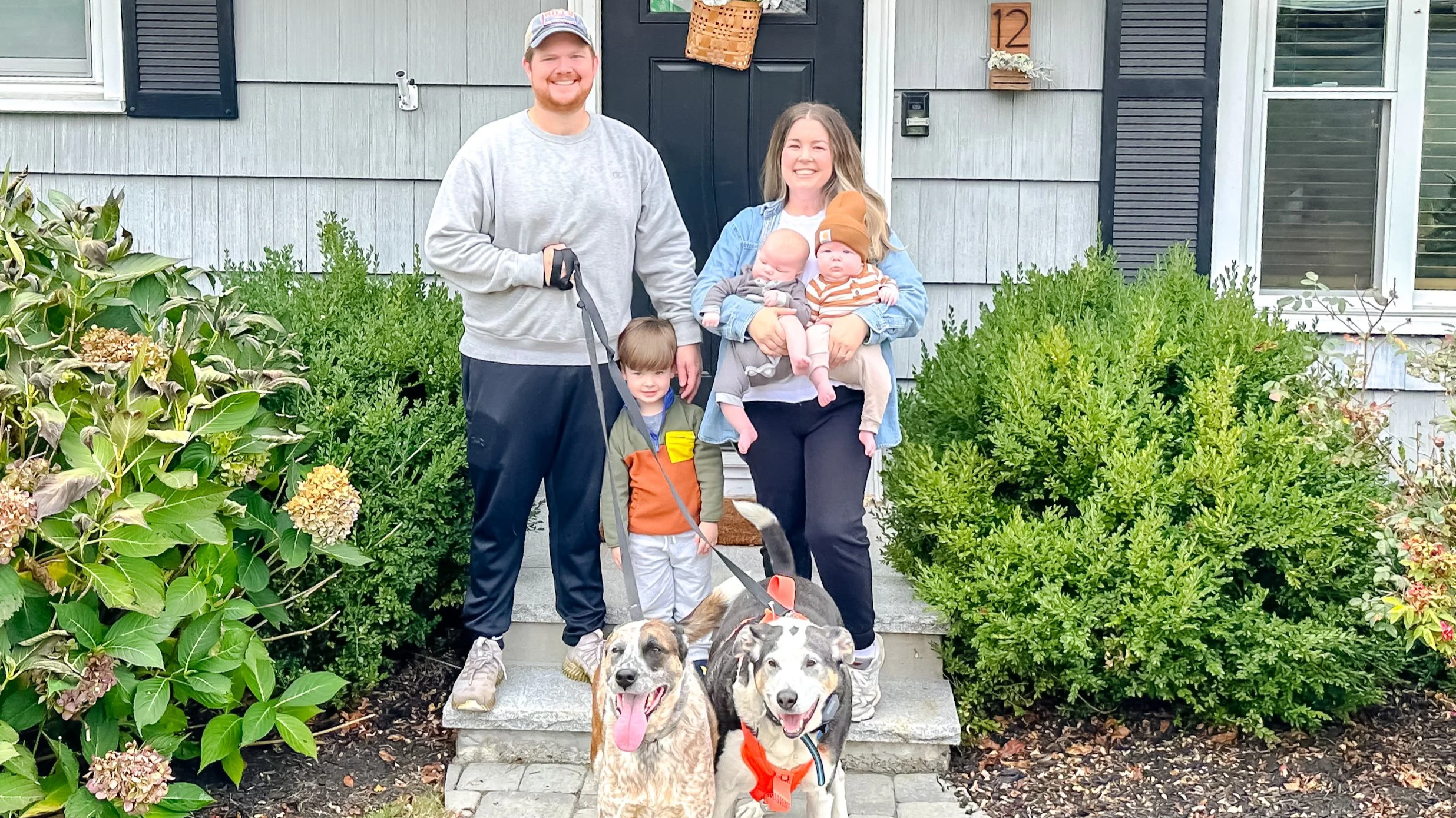 Family standing on front porch of house, with two dogs in front, surrounded by greenery.
