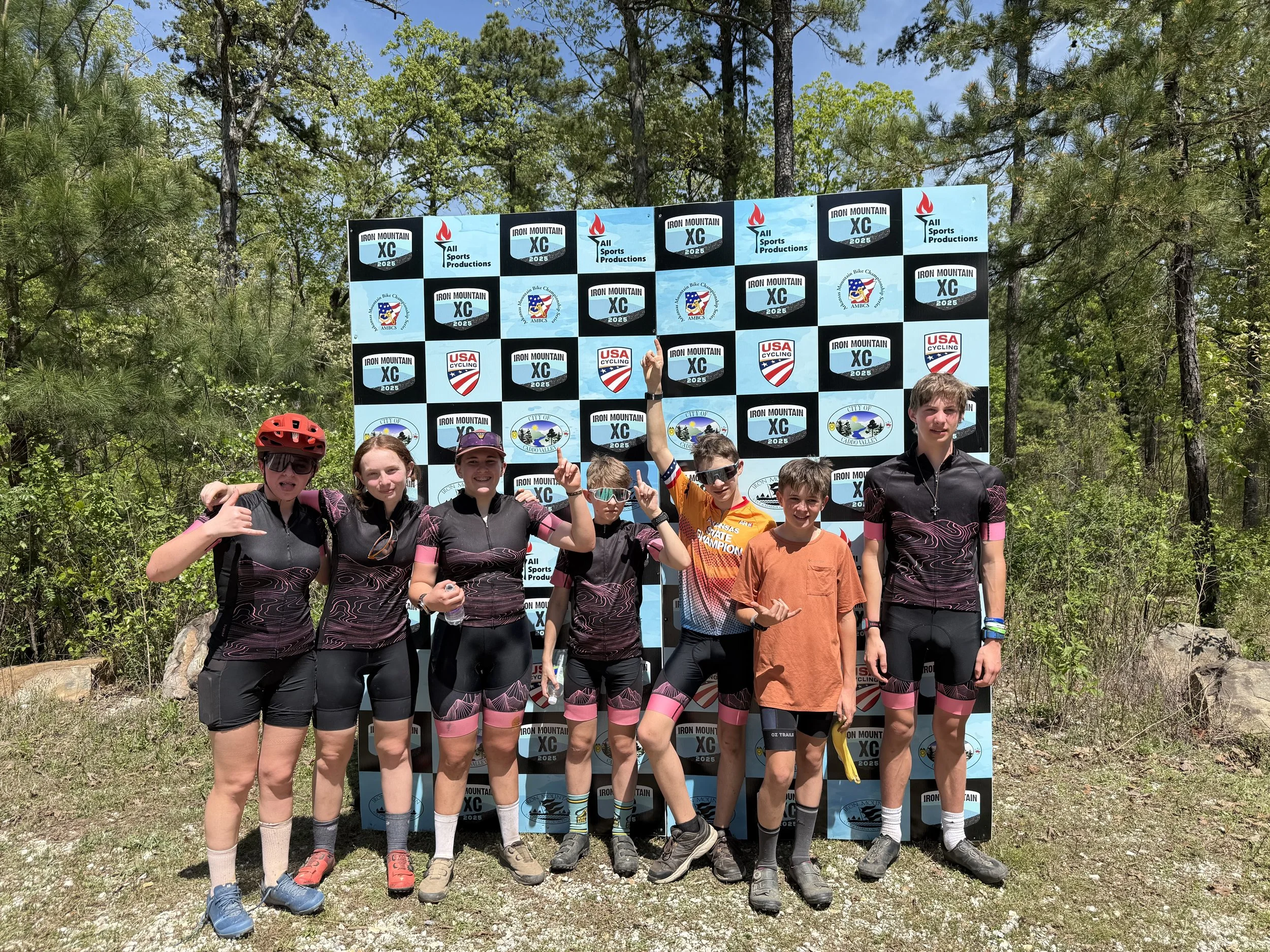 A group of seven children standing outdoors in front of a checkered backdrop with logos related to cross-country racing and cycling. The children are wearing sports attire, some with matching jerseys and shorts, and one child is wearing a cyclist's helmet. They are posing for a photo on a sunny day in a natural setting with trees and greenery in the background.