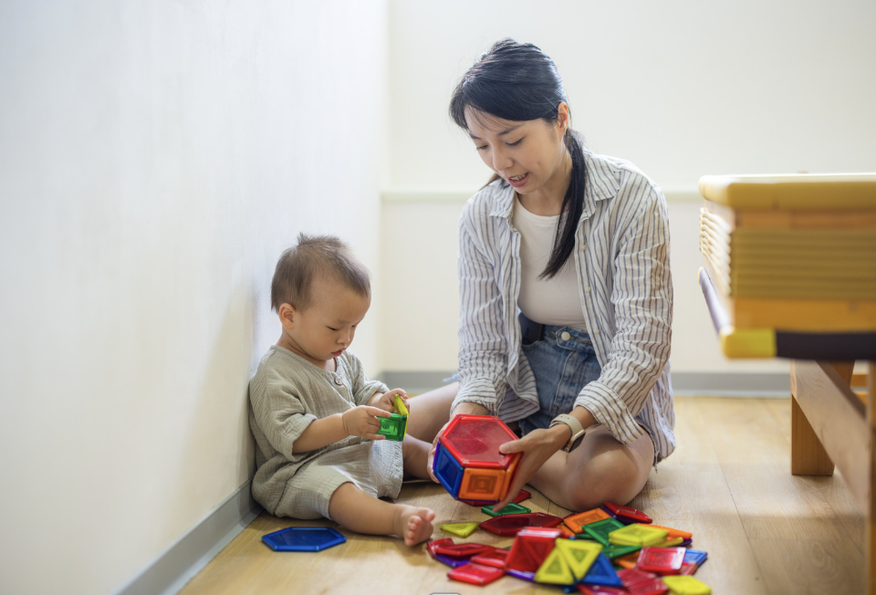 Woman and young child playing with colorful geometric shape toys on the floor in a bright room.