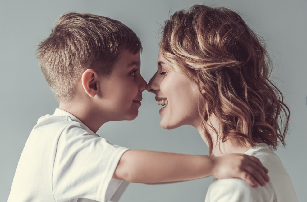 A young boy and a woman, likely his mother, face each other with their noses touching, smiling and showing affection.
