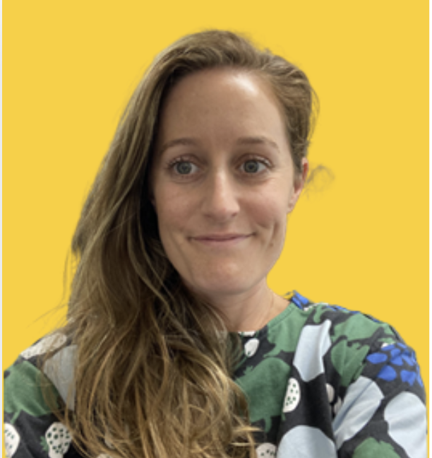 A woman with long, wavy, light brown hair smiling against a yellow background, wearing a multicolored patterned top.