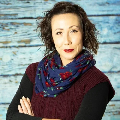 A woman with curly brown hair, wearing a burgundy sweater with black sleeves and a blue scarf with red floral patterns, standing outdoors in front of a weathered wooden background.