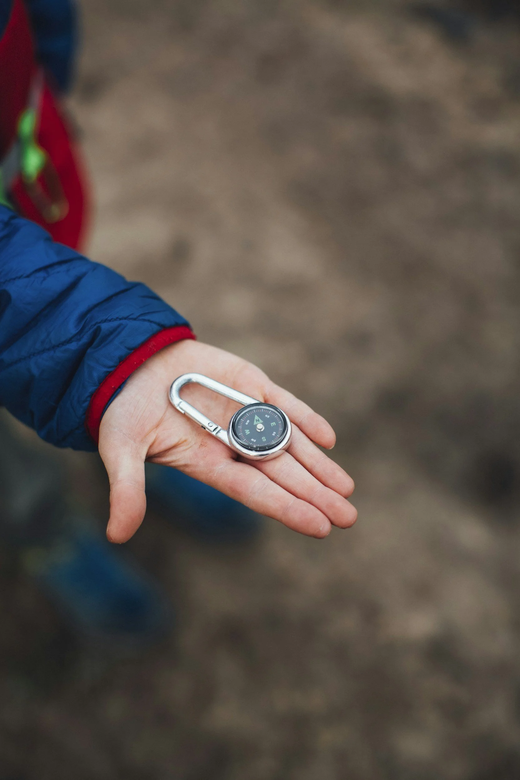 A person holding a silver carabiner with a built-in compass in their hand, wearing a blue jacket with red cuffs, standing on a dirt trail.