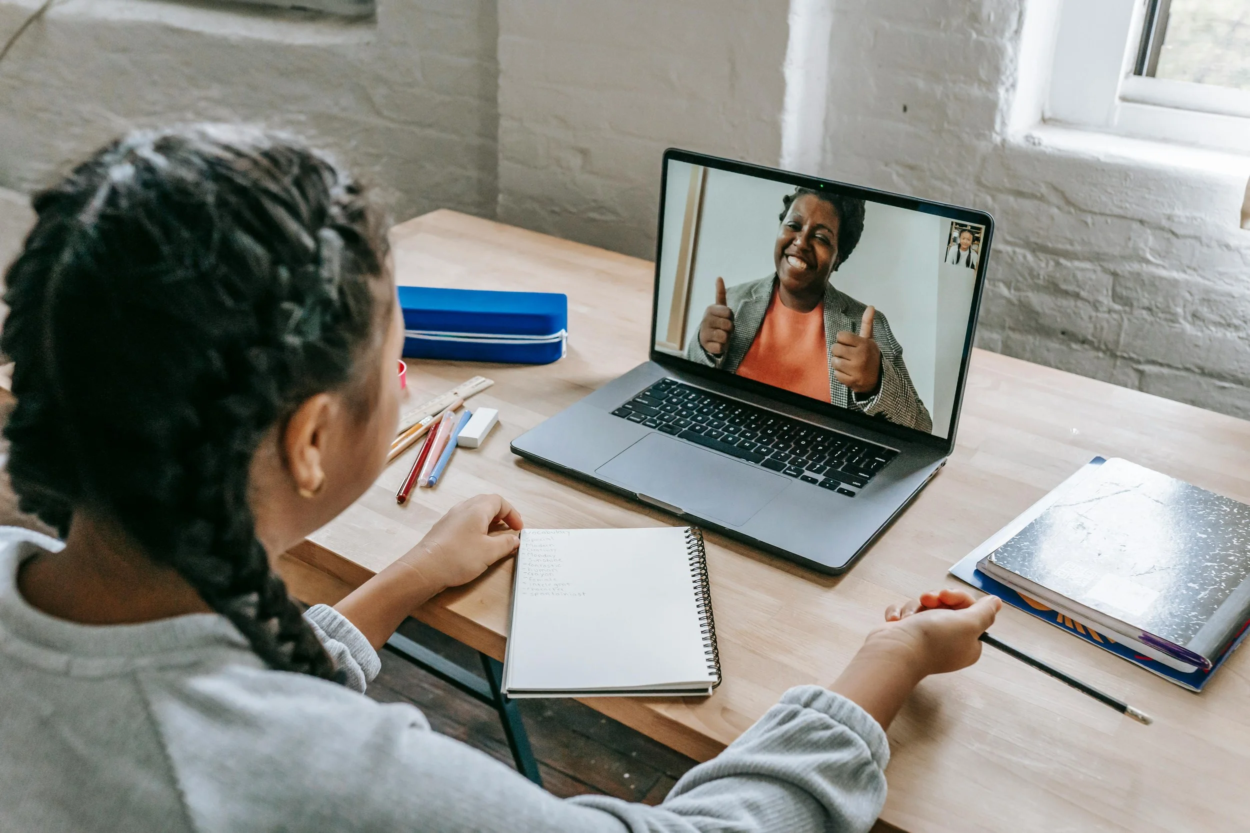 A girl with braided hair is sitting at a desk, having a video call on a laptop with a woman giving a thumbs up and smiling.