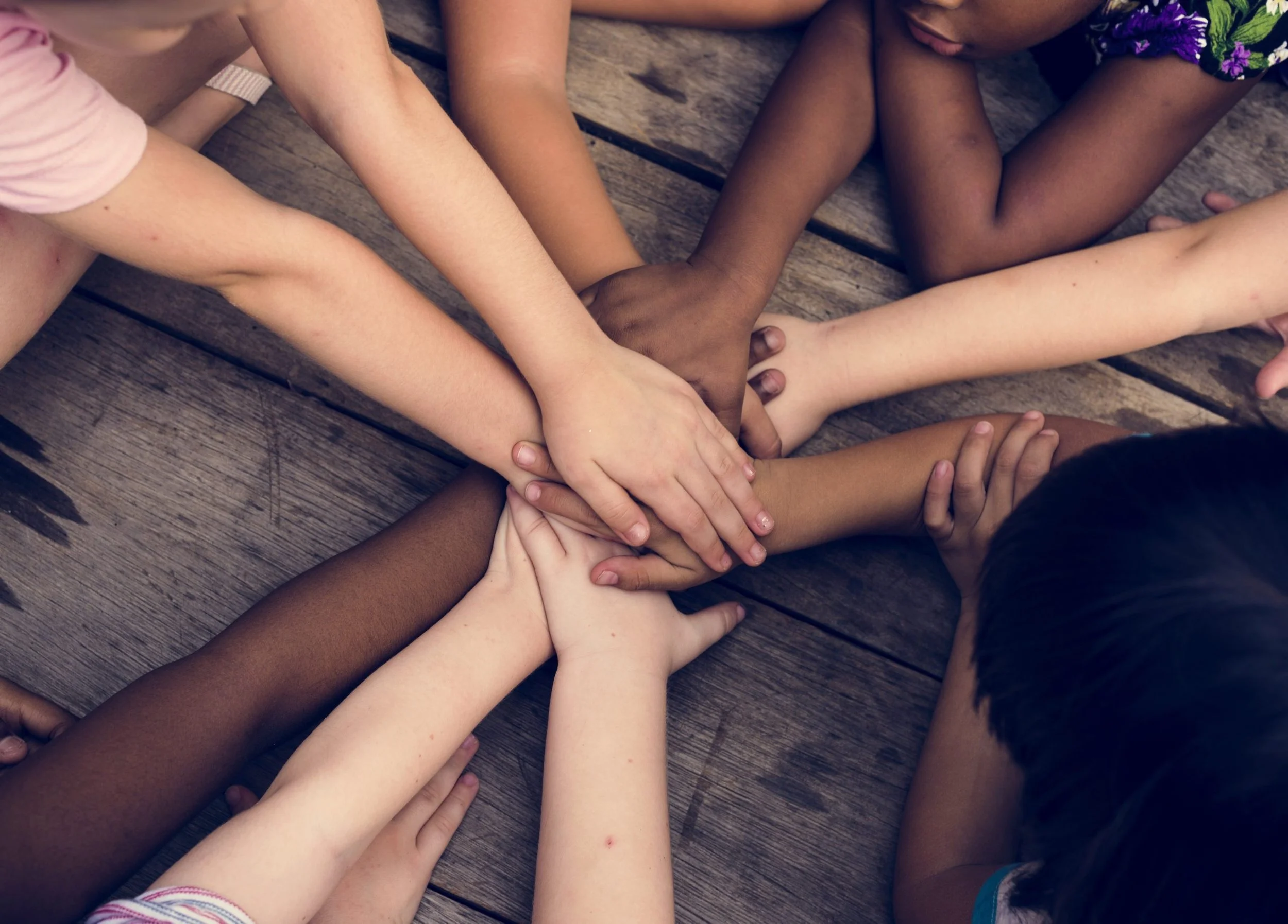 A group of children with different skin tones stacking their hands together in a circle on a wooden surface.