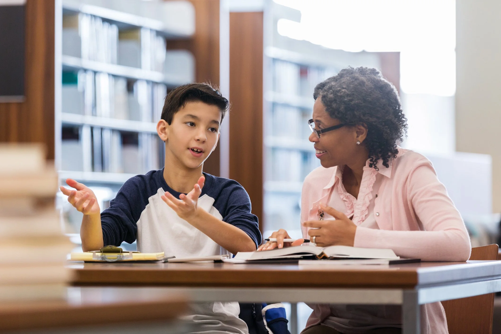 A woman and a young boy are sitting together at a table in a library, engaged in a conversation. The boy is gesturing with his hands while speaking, and the woman is listening attentively. There are books and notepads on the table.