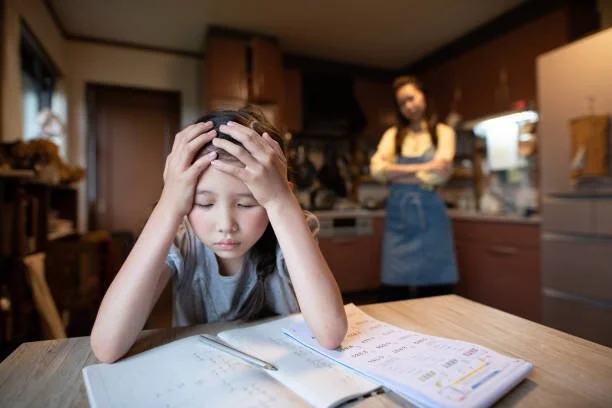 A young girl sitting at a table with a distressed expression, holding her head with both hands, in a kitchen. An adult woman is standing in the background, overseeing from a distance.