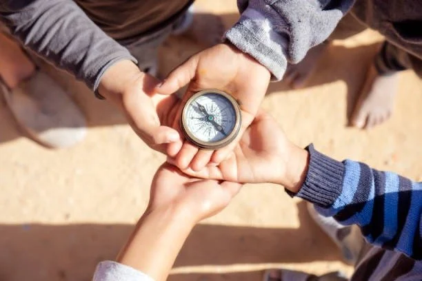 Four hands overlapping, holding a compass in the center, viewed from above outdoors on sandy ground.