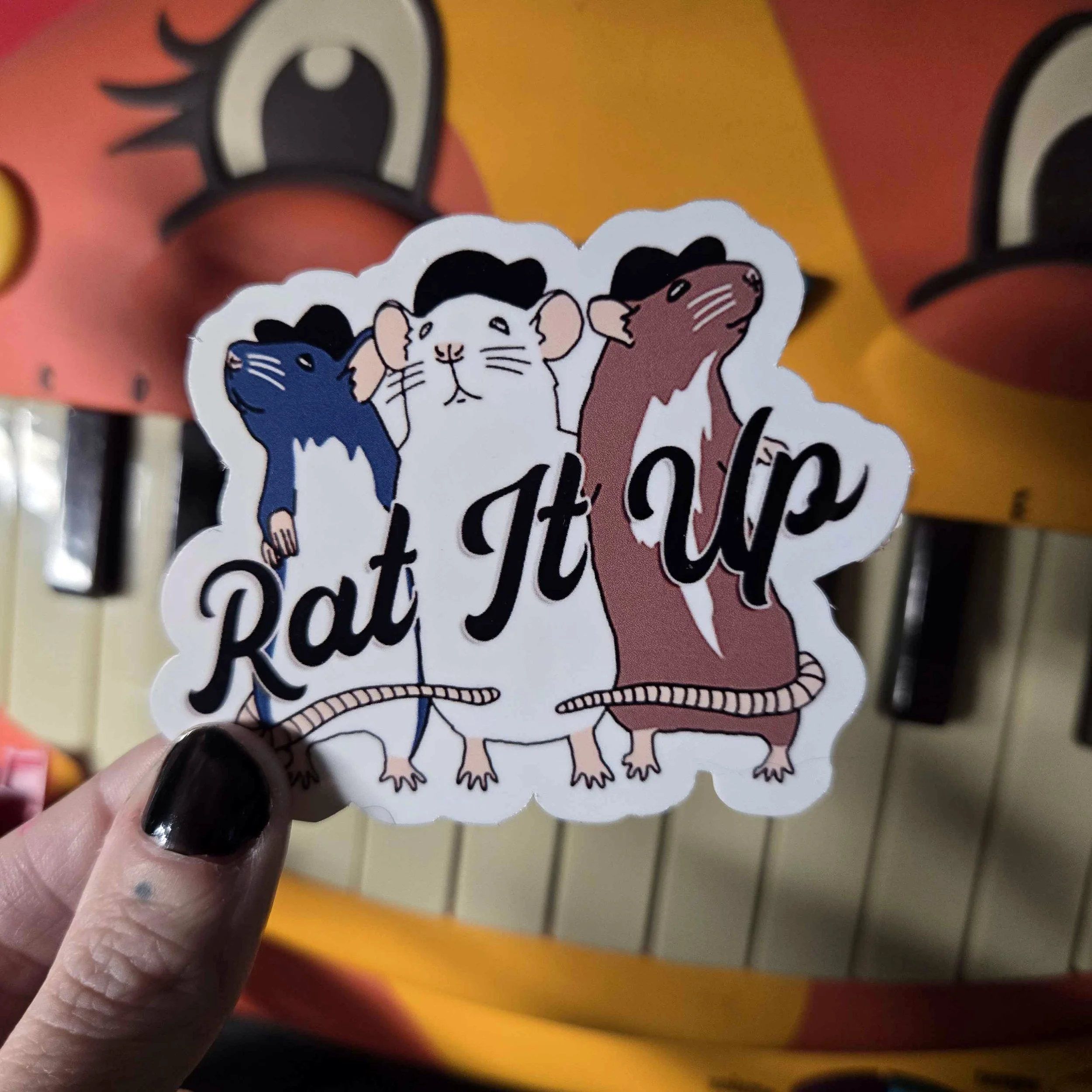 a hand holding a rat sticker of three rats with their arms around each other standing in a row all wearing black hats, and the words "rat it up" over their chests written in black. the background is of an orange cat keyboard.