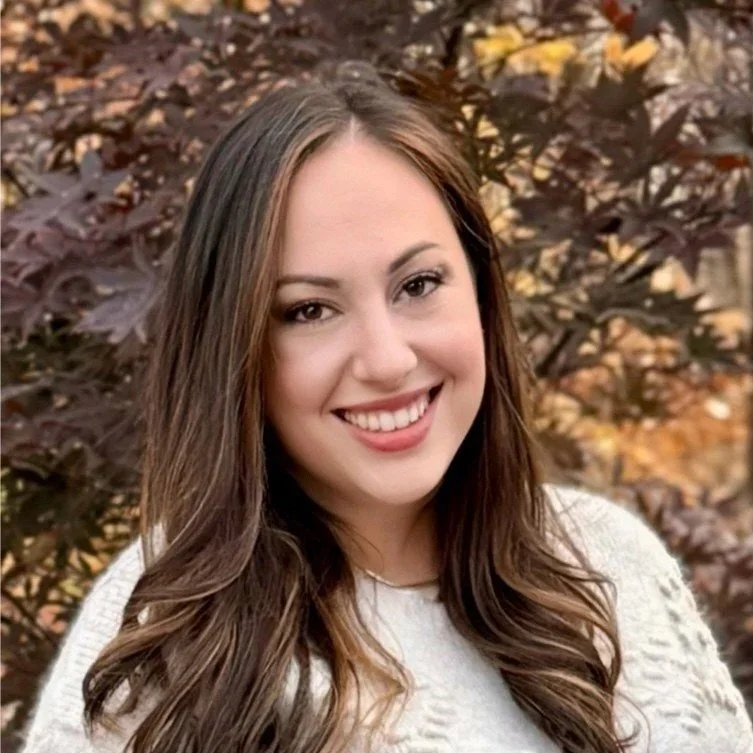 A woman with long brown hair and a white top smiling outdoors with autumn leaves in the background.