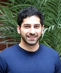 A young man with dark curly hair and a beard, smiling, wearing a navy blue shirt, standing outdoors with green plants in the background.