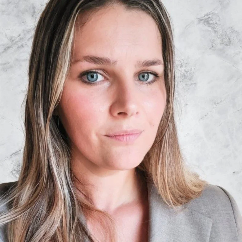 Close-up portrait of a woman with shoulder-length light brown hair, blue eyes, and fair skin, wearing a gray blazer, standing in front of a light gray textured wall.
