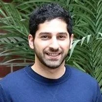 A smiling man with dark curly hair and a beard, wearing a blue shirt, standing outdoors with green foliage in the background.