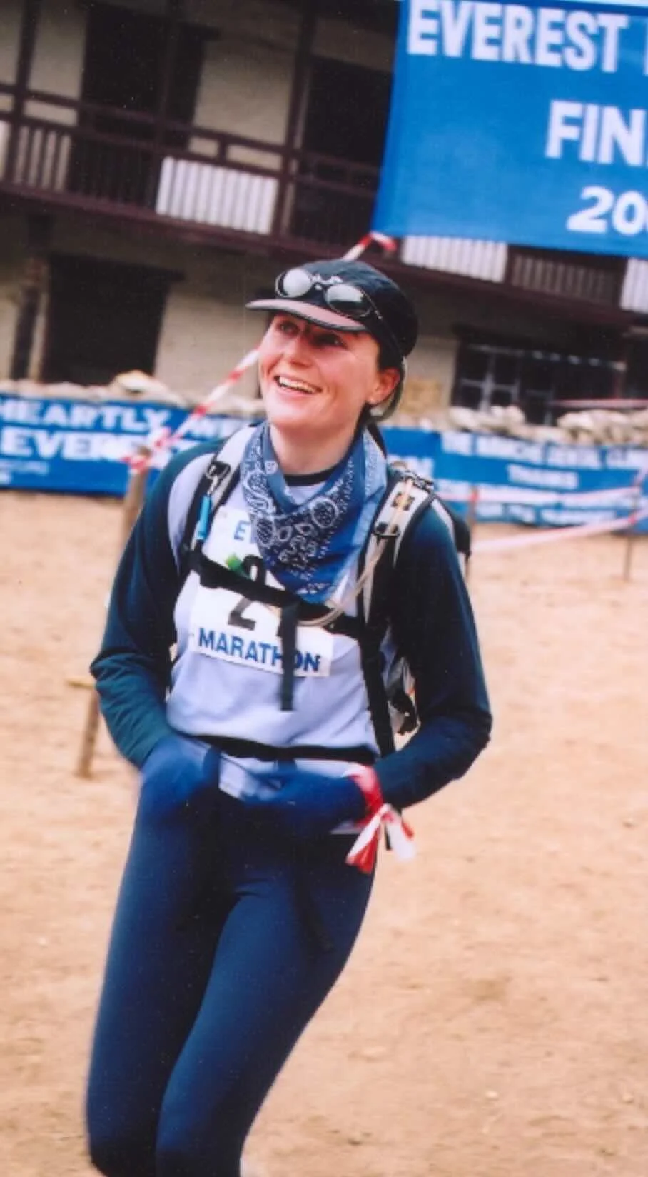 Female marathon runner smiling, wearing a black cap with goggles, blue bandana around neck, navy jacket, and black leggings, participating in a marathon event at Everest finish line.