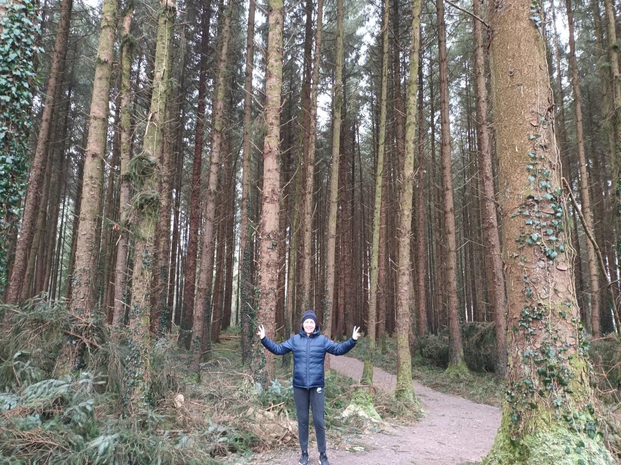 A person standing on a forest trail surrounded by tall trees with a joyful expression, making peace signs with both hands.