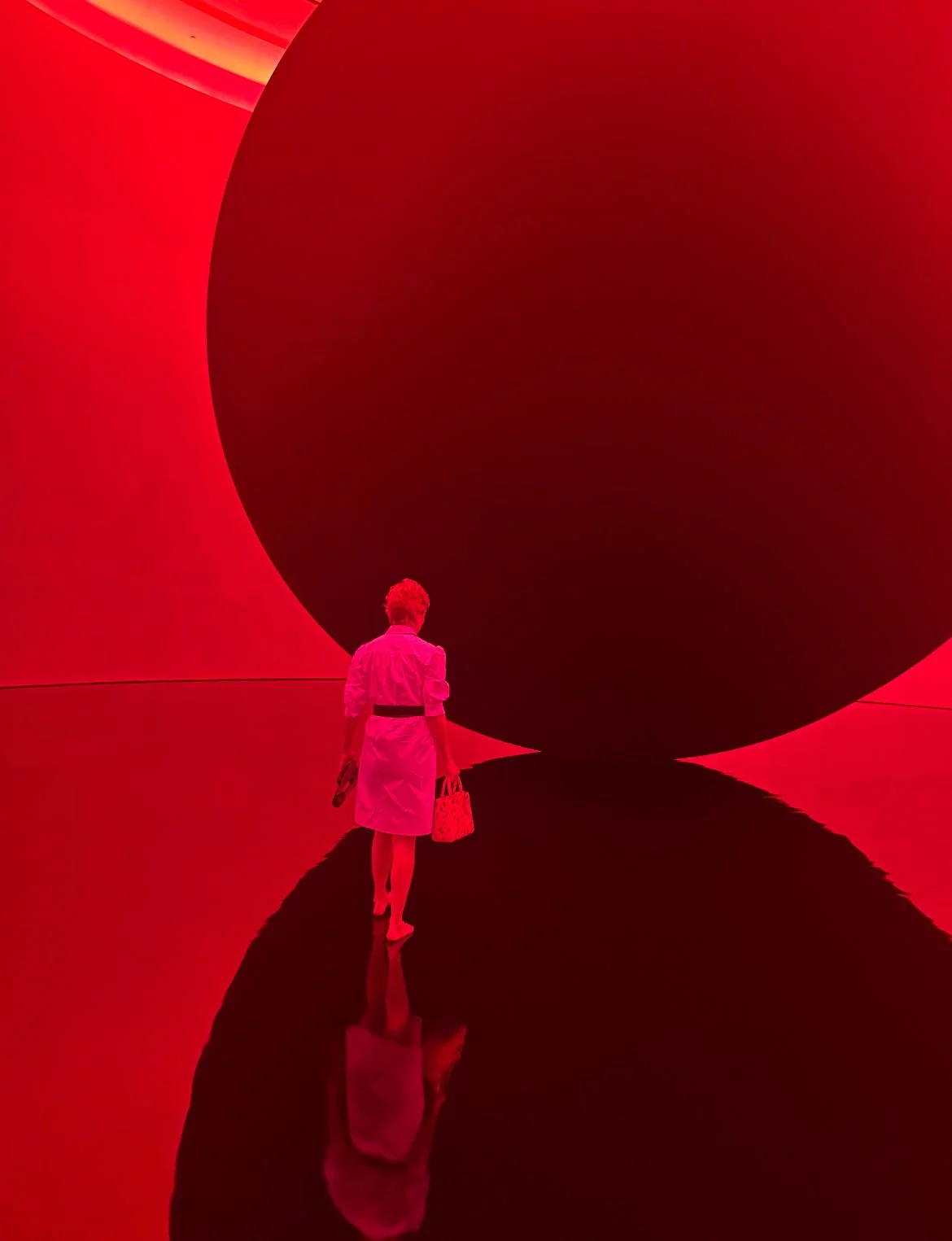 A woman with blonde hair in a white coat holding a patterned bag and a phone walking on a reflective black floor, surrounded by red glowing walls and large abstract shapes.