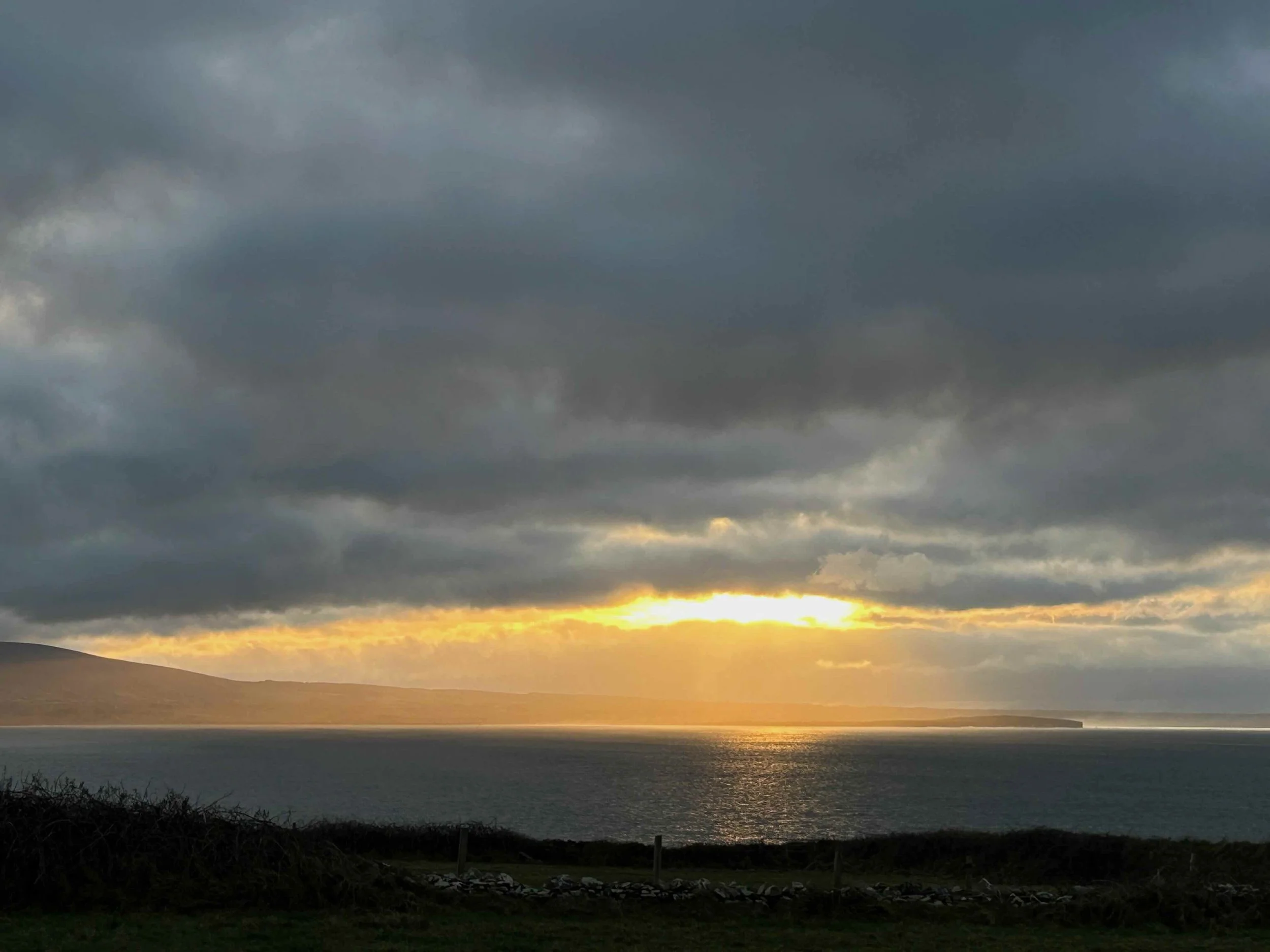 Sea with a distant headland at sunset, dark clouds overhead, and light breaking through clouds reflected on the water.