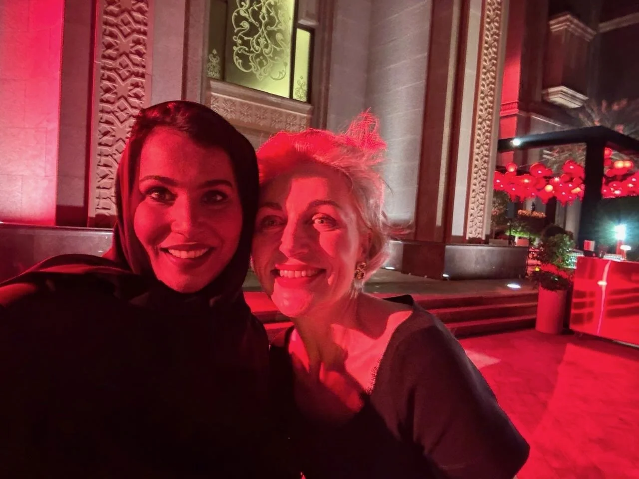 Two women smiling and taking a selfie in front of a decorative building entrance illuminated with red lighting, with a display of red lanterns or ornaments hanging in the background.