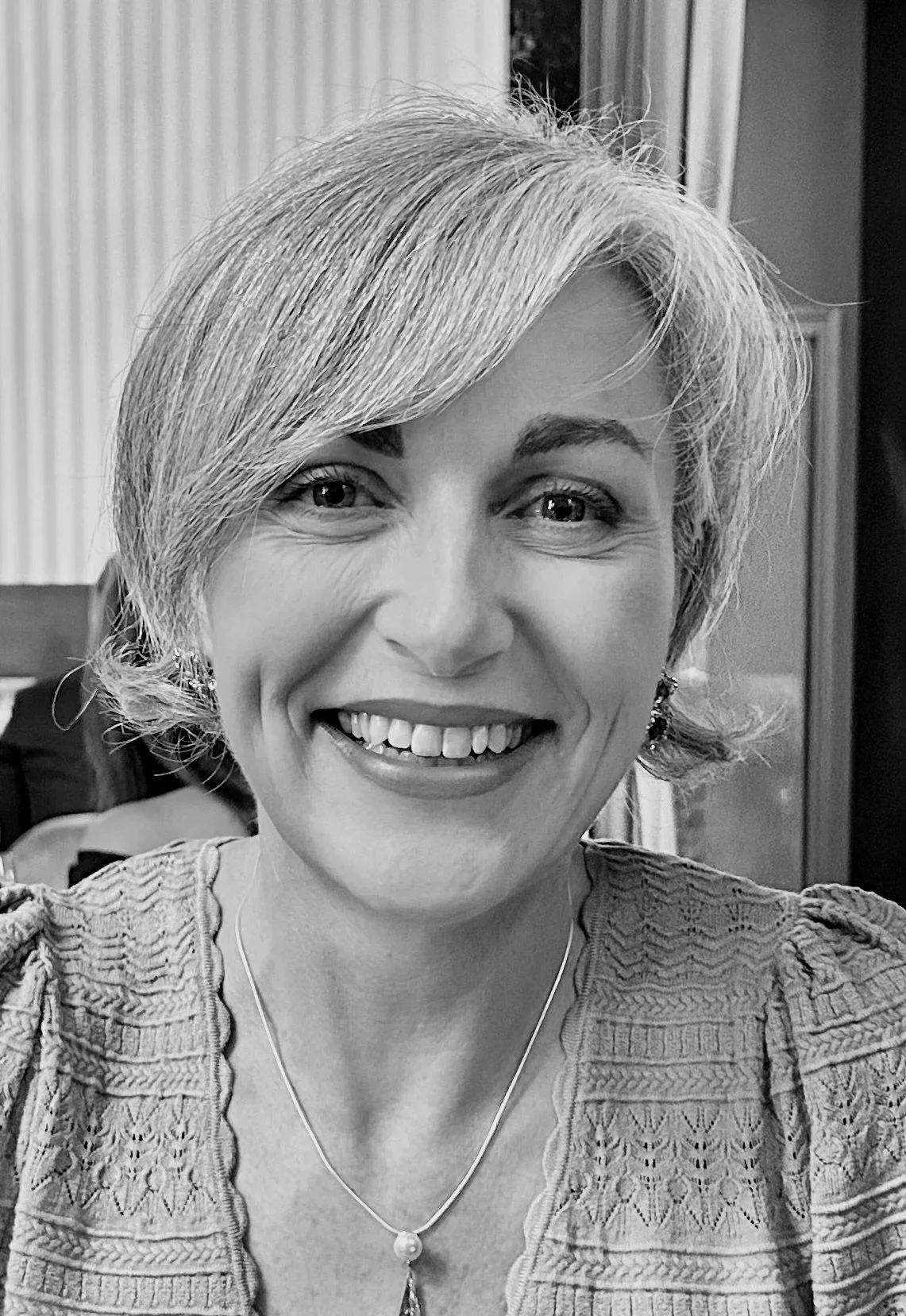 A smiling woman with short gray hair, wearing earrings and a necklace with a pendant, in a black and white photo.