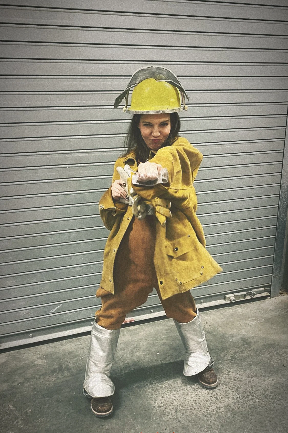 Woman dressed in bronze casting gear wearing a yellow helmet, yellow jacket, brown pants, and silver boot coverings, standing in front of a metal garage door, pointing forward with a confident expression.