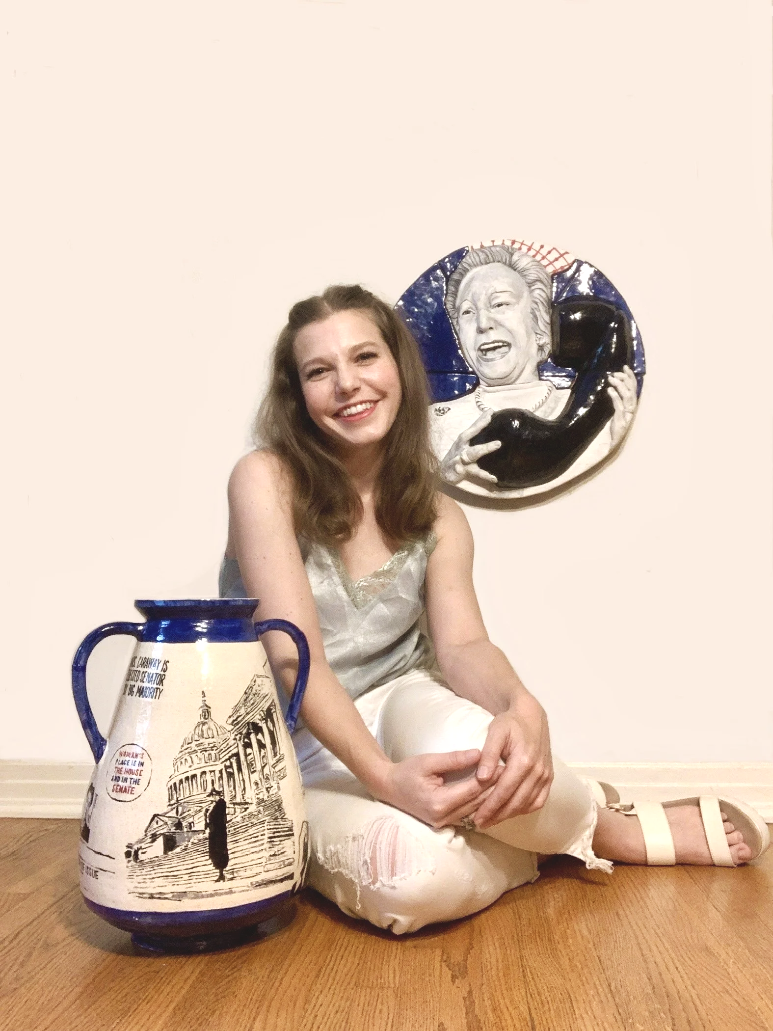 A smiling woman with long brown hair, sitting on the floor, poses next to a large ceramic pot and a ceramic relief of a woman holding a telephone, both with blue accents, against a plain white wall.
