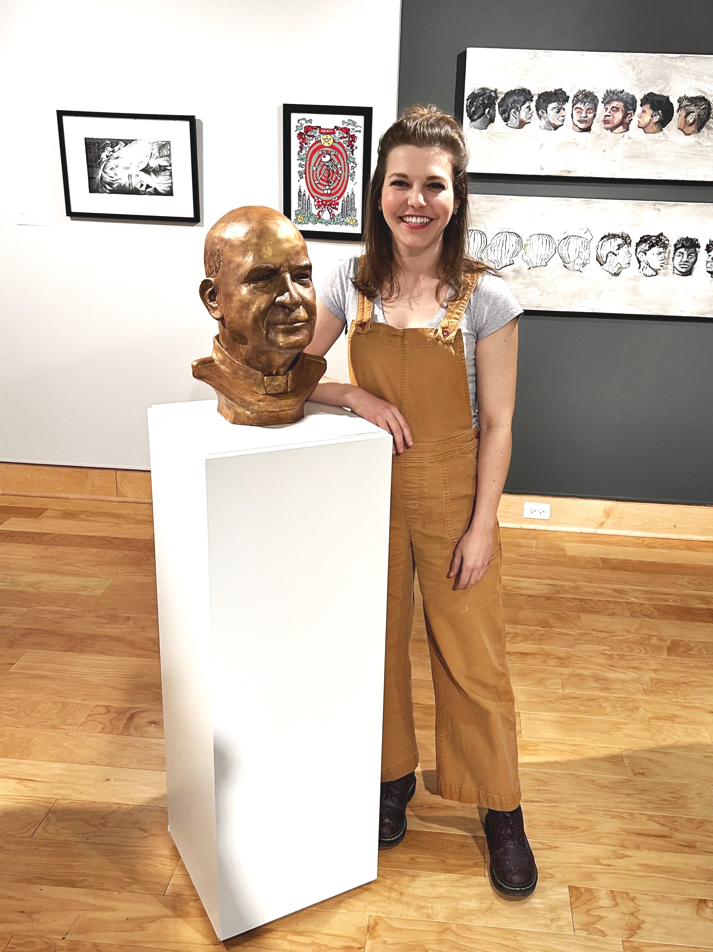 A woman with brown hair and a big smile is standing beside a bronze bust of a man on a white pedestal.