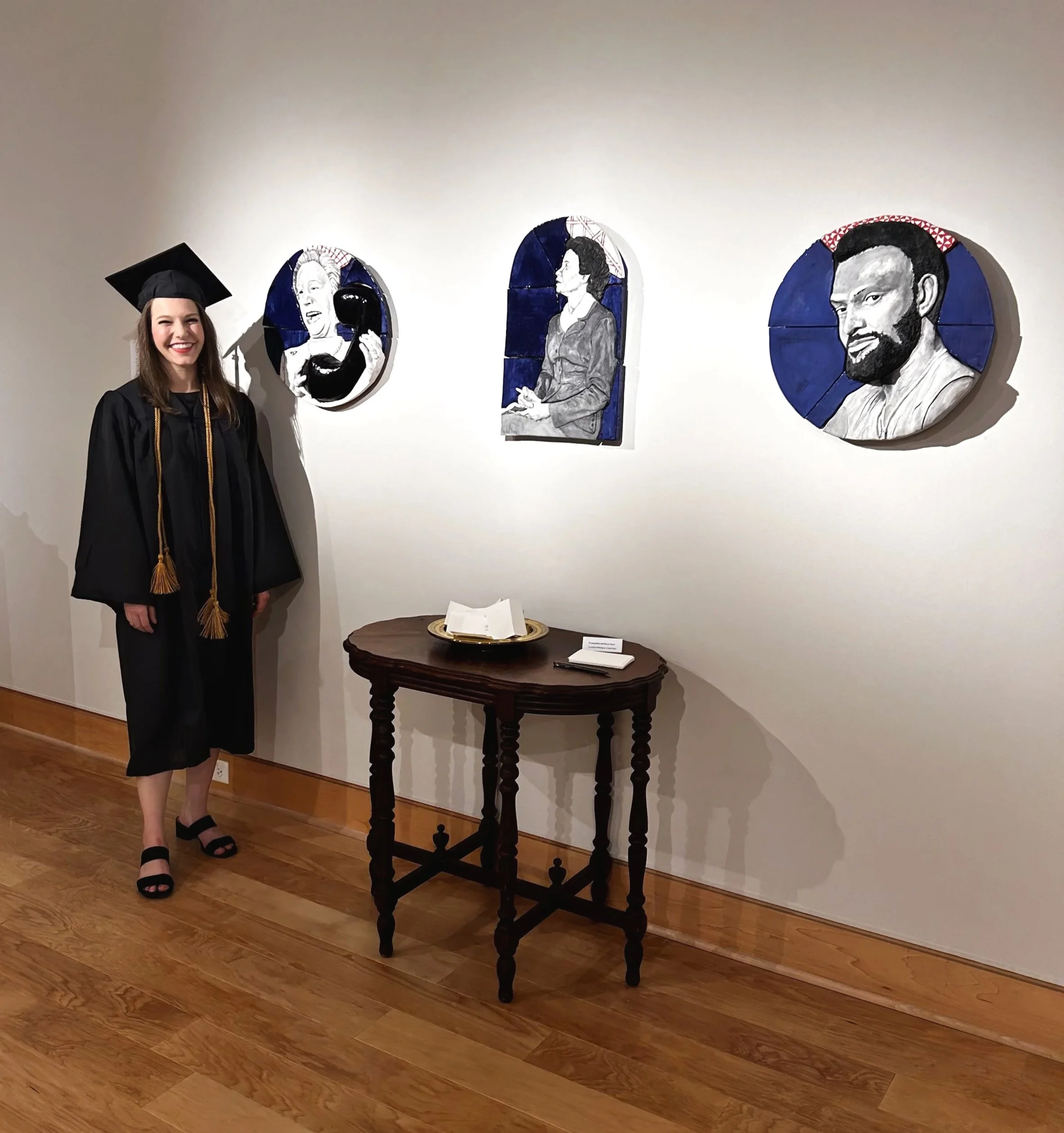 A young woman in a black graduation gown and cap with a gold tassel stands in an art gallery, smiling at camera. Behind her are three circular art pieces on the wall with black-and-white portraits of people, each framed with dark blue backgrounds.