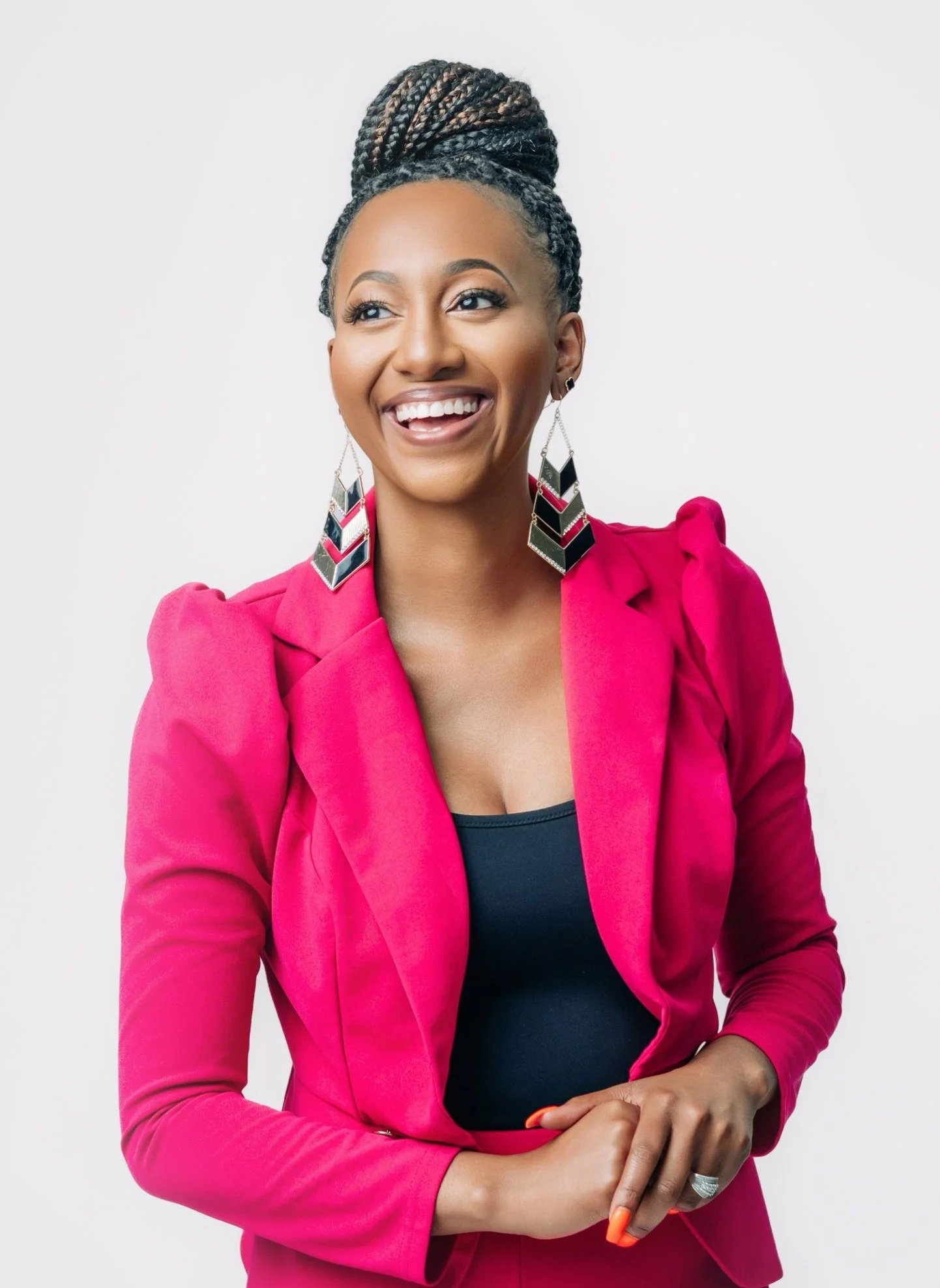 A woman with braided hair styled in an updo, wearing a bright pink blazer over a black top, with large geometric earrings, smiling with her hands clasped in front of her against a plain white background.