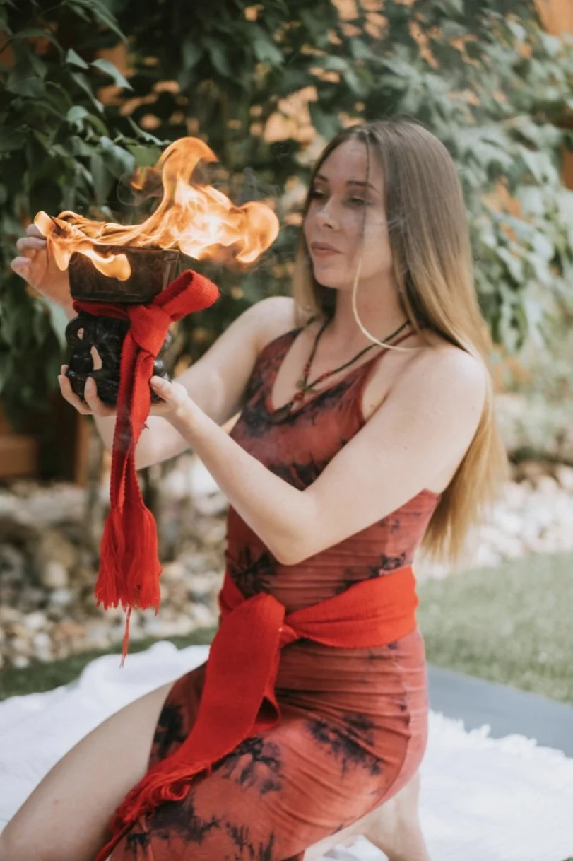 A woman in a red tie-dye dress holding a flaming fire bowl outdoors.