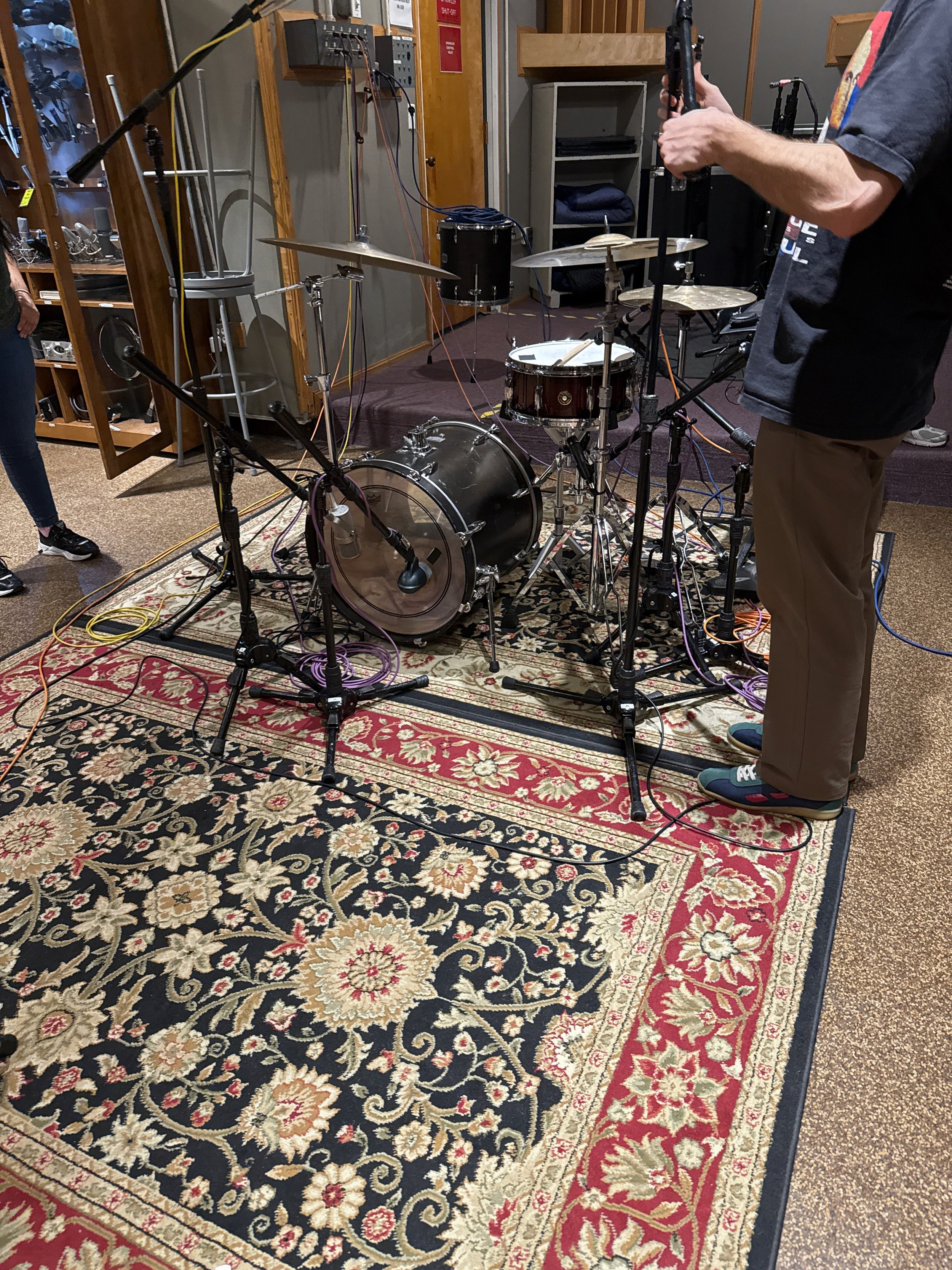 A drum set on an ornate patterned rug in a recording studio, with multiple microphones and audio equipment around it. A person is standing nearby holding a guitar or similar instrument.