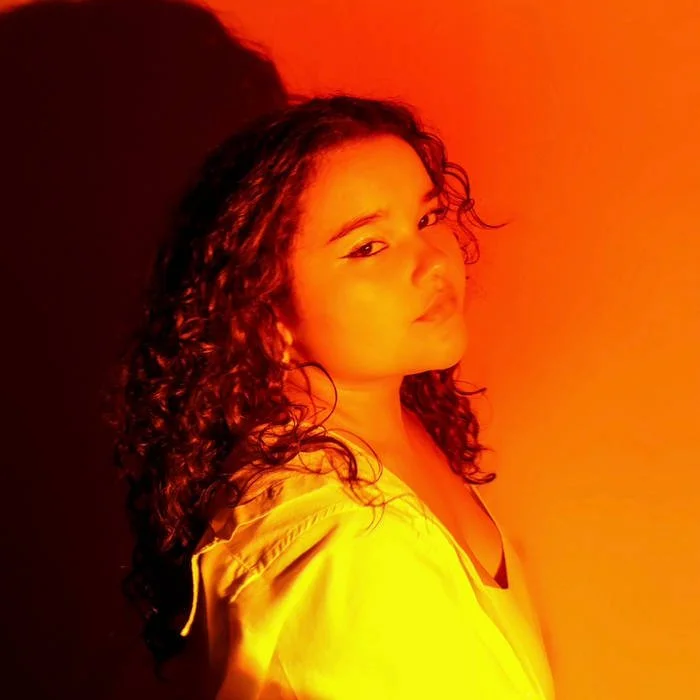 A woman with curly hair posing against a warm, orange-toned background, wearing a yellow jacket.