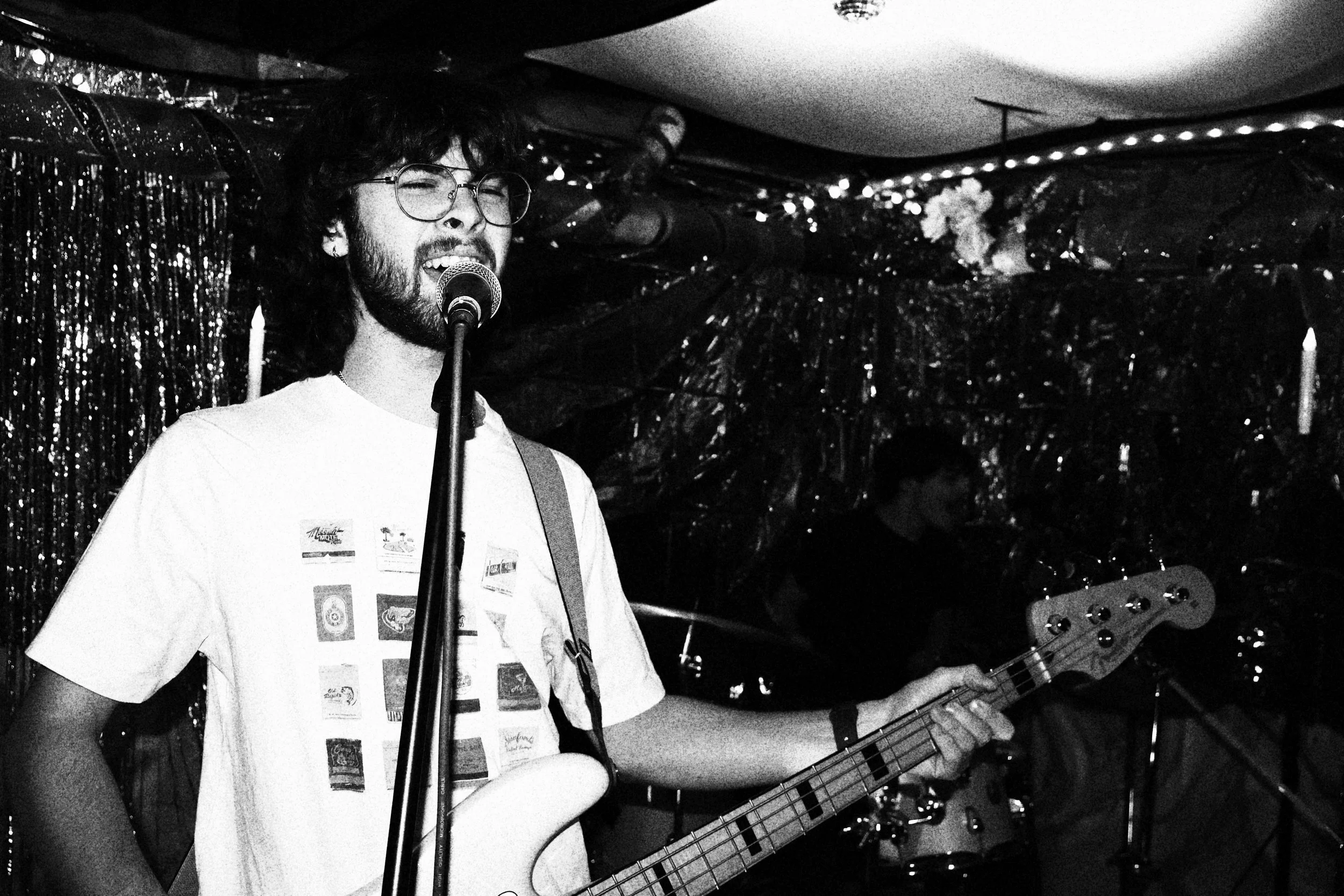 A young man with glasses and a beard singing into a microphone while playing an electric bass guitar at a performance, with another musician visible in the background.