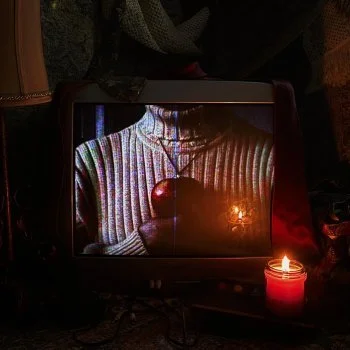A small candle in a red holder, a person wearing a striped sweater with a pendant necklace, and a reflection of a lit candle on a television screen.