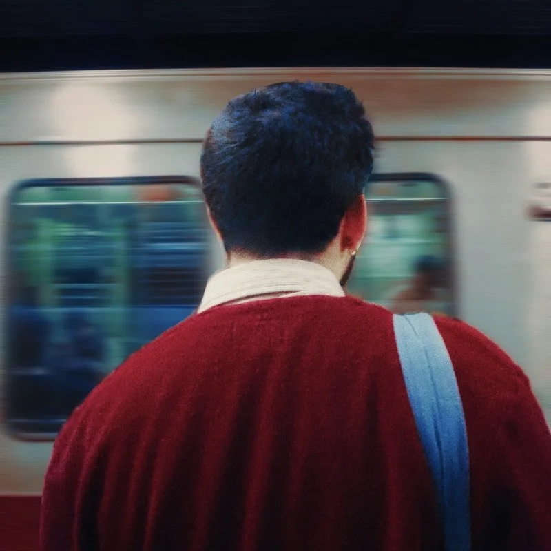 A person with short, dark, curly hair wearing a red jacket and carrying a gray strap backpack, standing on a subway platform with a moving train behind them.