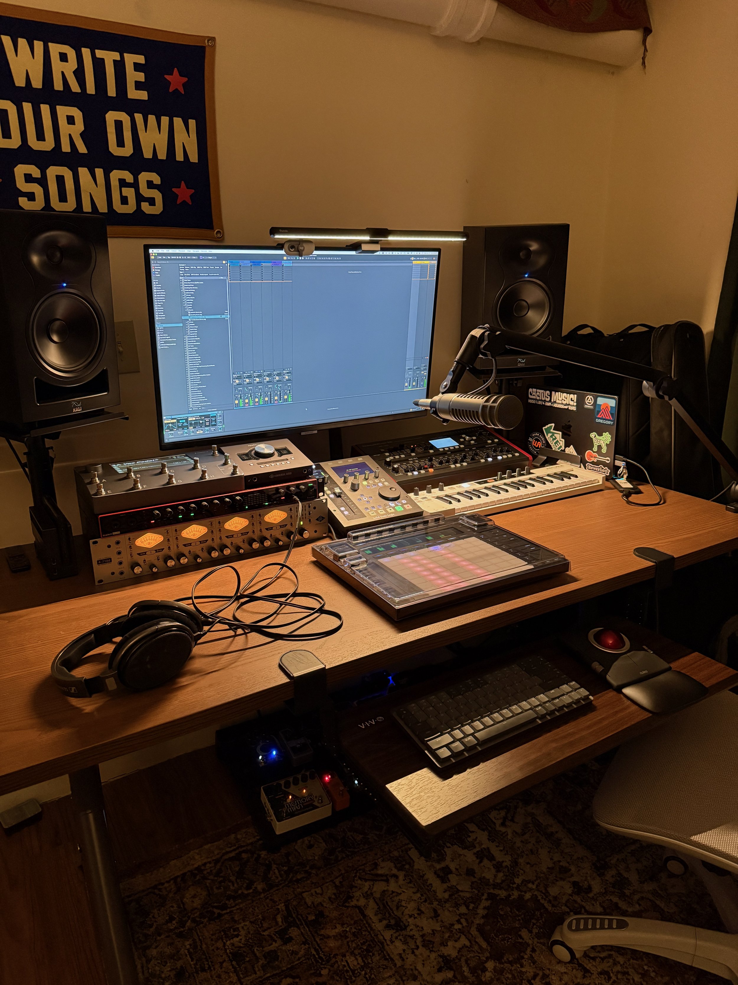 A home music studio setup featuring a large computer monitor, studio speakers, a microphone on a boom arm, a MIDI controller, audio interface, headphones, and various music production equipment on a wooden desk, with a sign on the wall that reads "WRITE YOUR OWN SONGS."