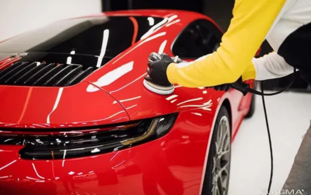 Person polishing a red sports car with a power buffer in a clean, well-lit environment.