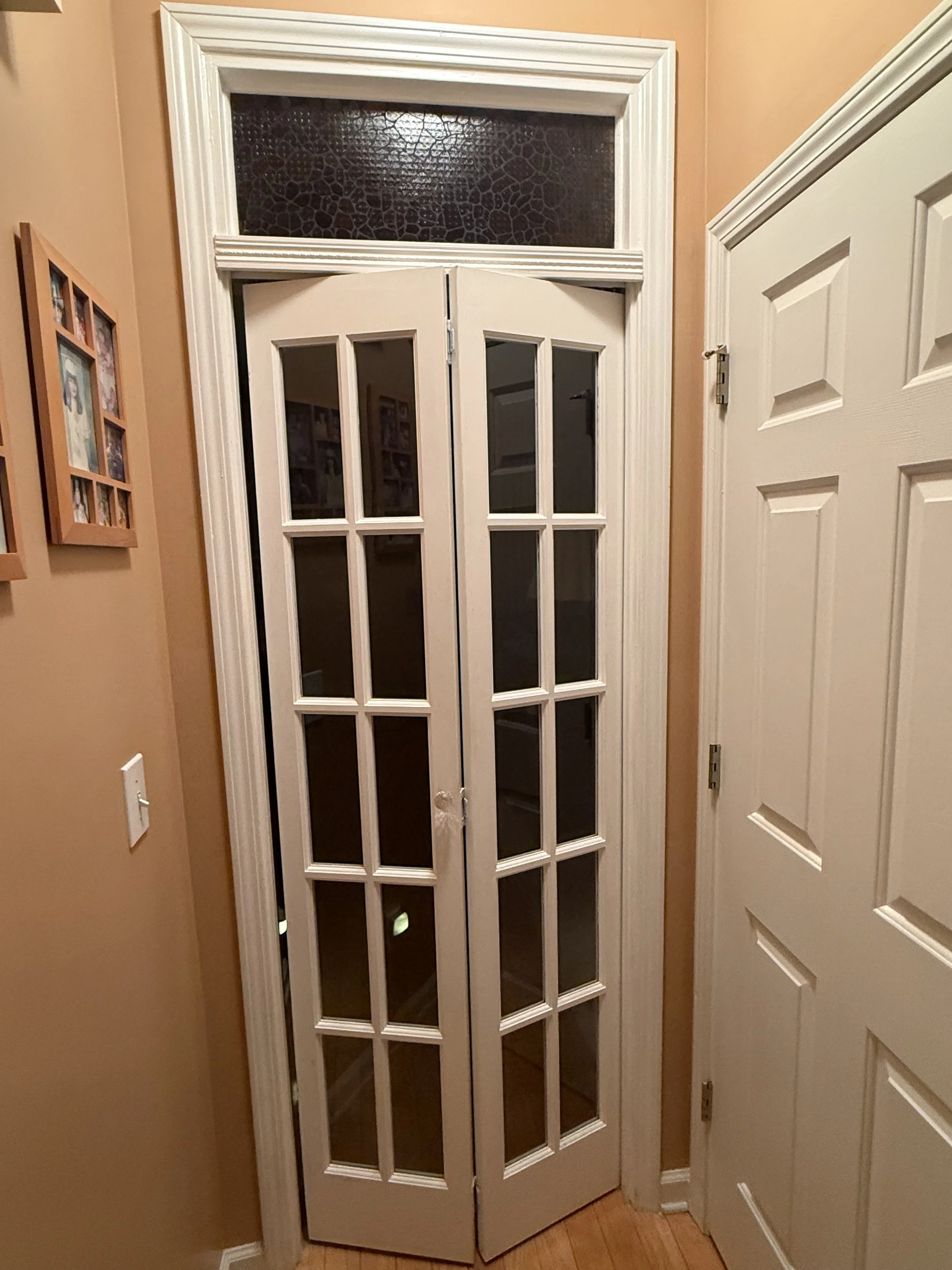 A set of glass-paneled double doors with white framing, leading into a room. The doors are slightly open, revealing a dark interior. The wall is painted a warm beige, and there is a framed photo collage on the left wall.
