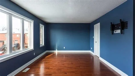 Empty room with blue walls, hardwood floor, three windows, white door, and wall-mounted TV bracket.