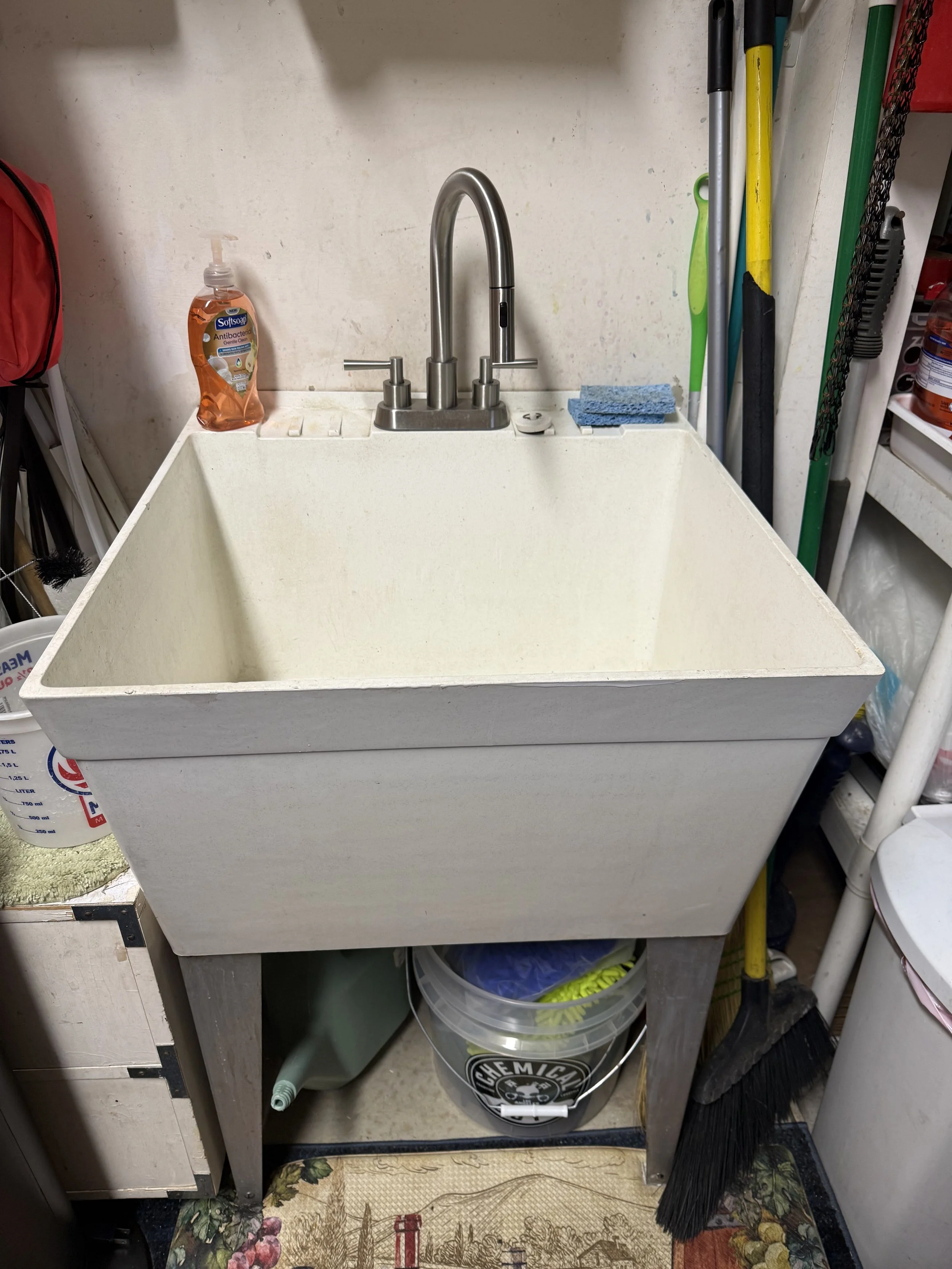 A utility or laundry room sink with a silver faucet. To the left, there is a bottle of antibacterial hand soap. To the right, cleaning tools such as a broom, mop, and squeegee are stored against the wall. The sink is on a metal stand, and a bucket with a chalk logo is underneath. The floor has a patterned rug.