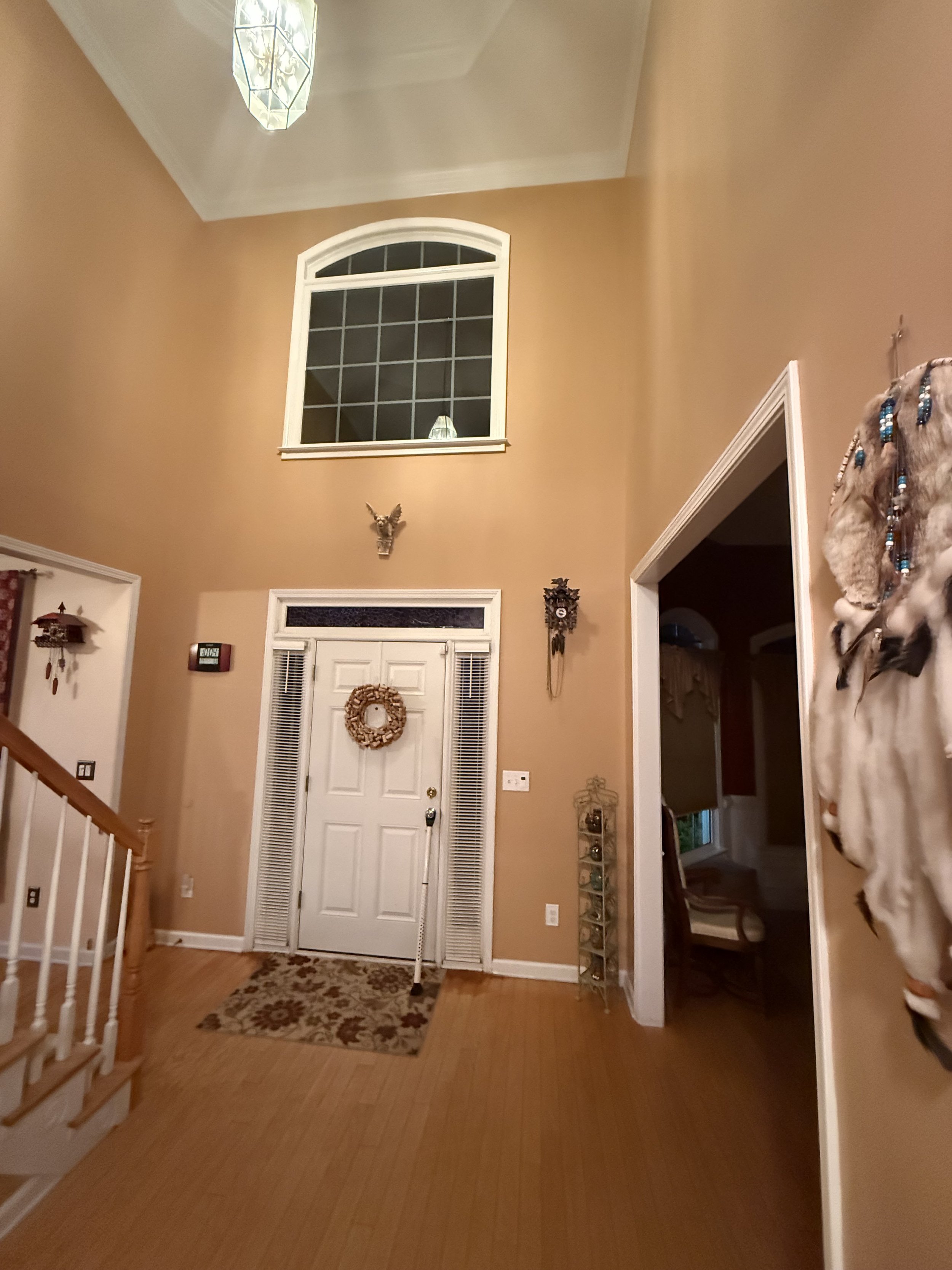 Entryway with a white front door, decorative wreath, and a small doormat. High ceiling with a window near the ceiling and a hanging light fixture. Warm-colored walls with wall decor, a staircase on the left, and an open doorway to other rooms.