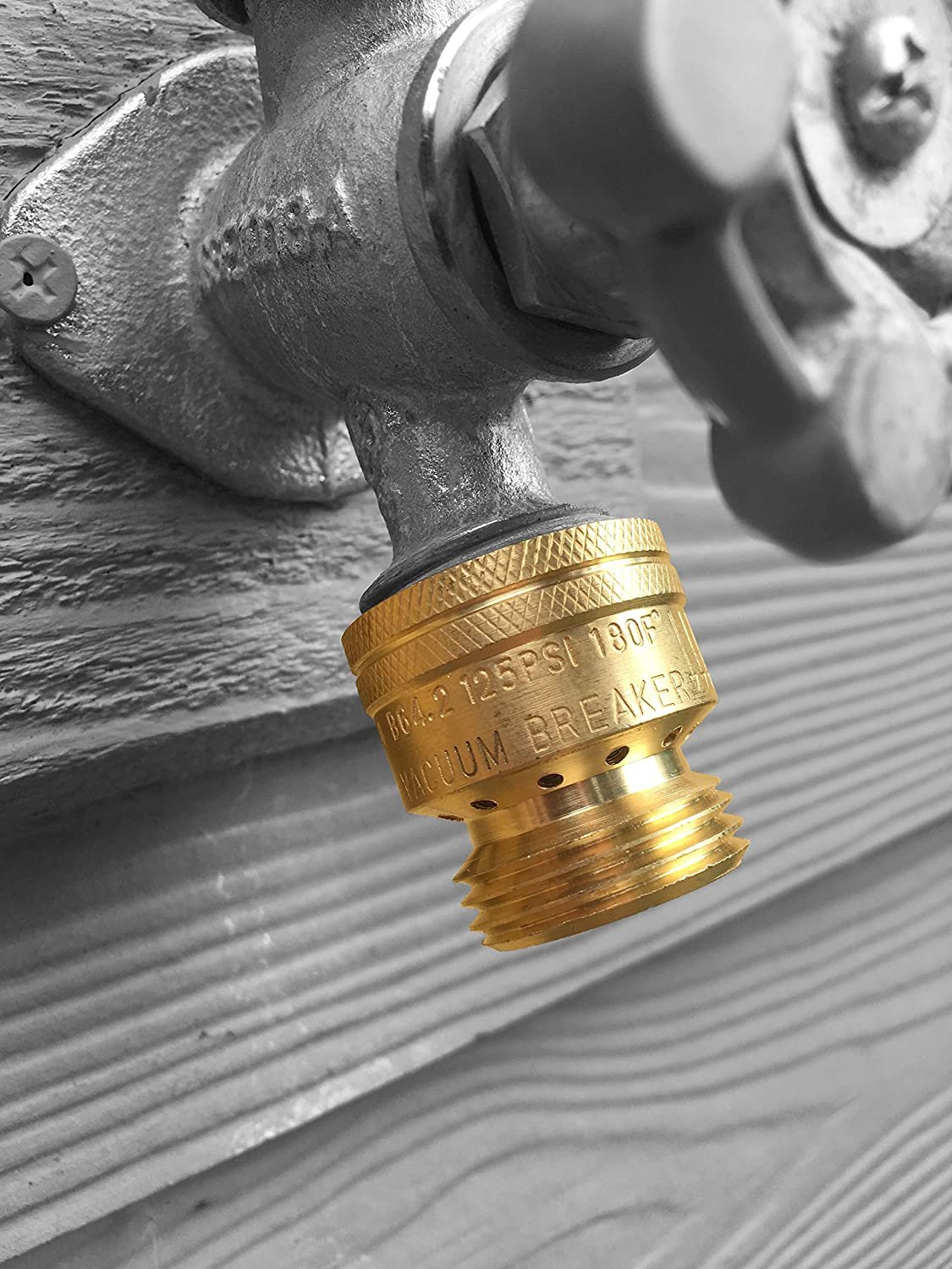 Close-up of a brass vacuum breaker fitting attached to a water pipe with a gray textured surface in the background.