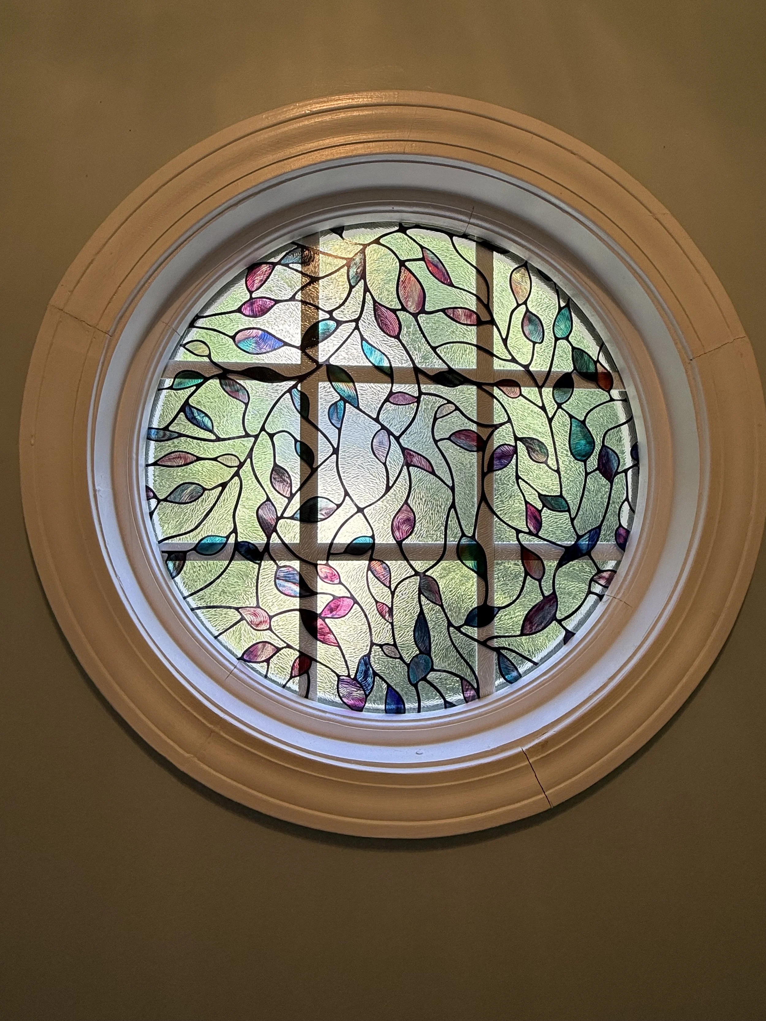 Round stained glass window with a colorful leaf pattern, set in a white frame against a beige wall.