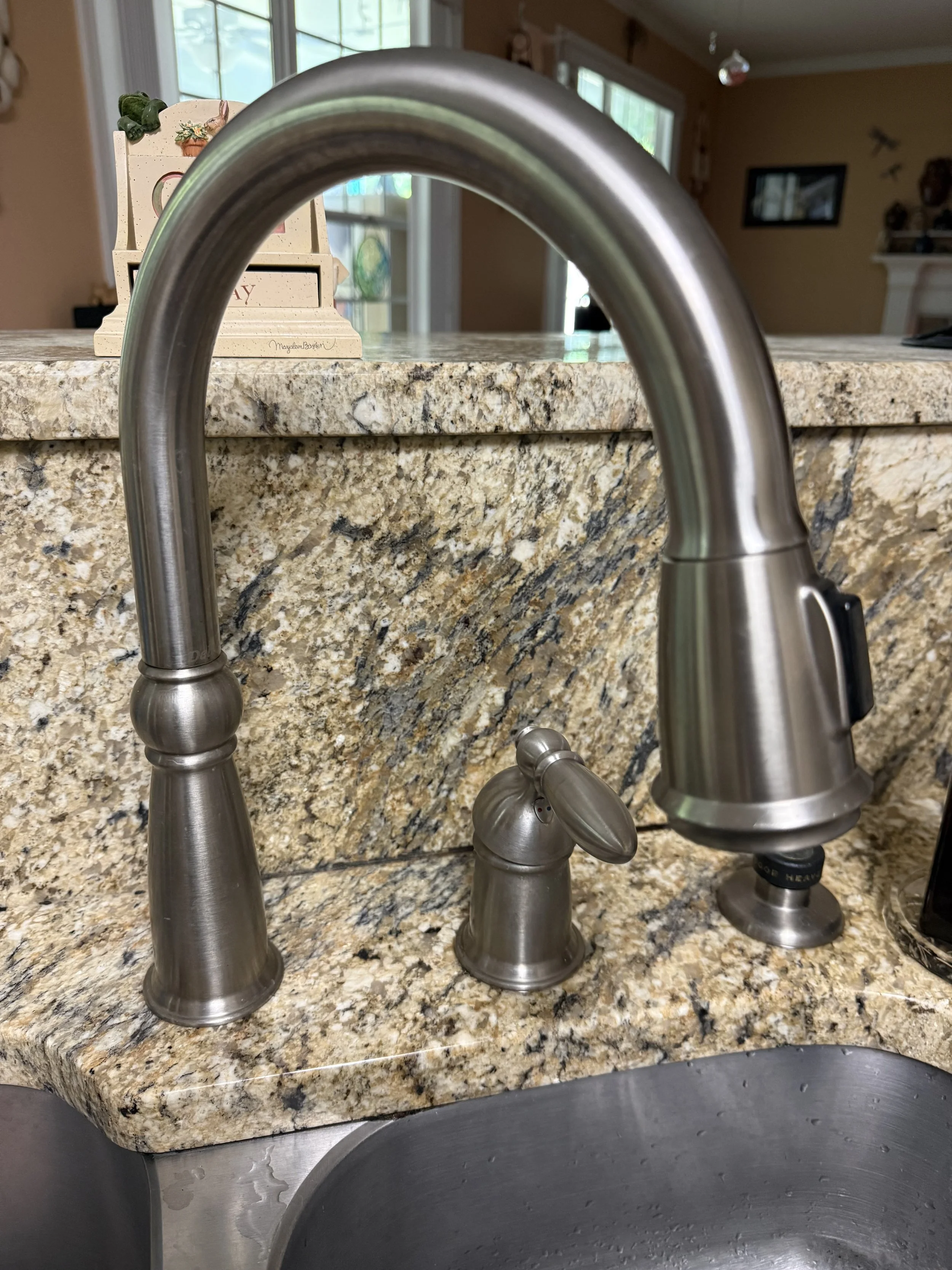 Close-up of a kitchen sink with a stainless steel faucet, granite countertop, and background featuring a decorative wooden box and kitchen window.
