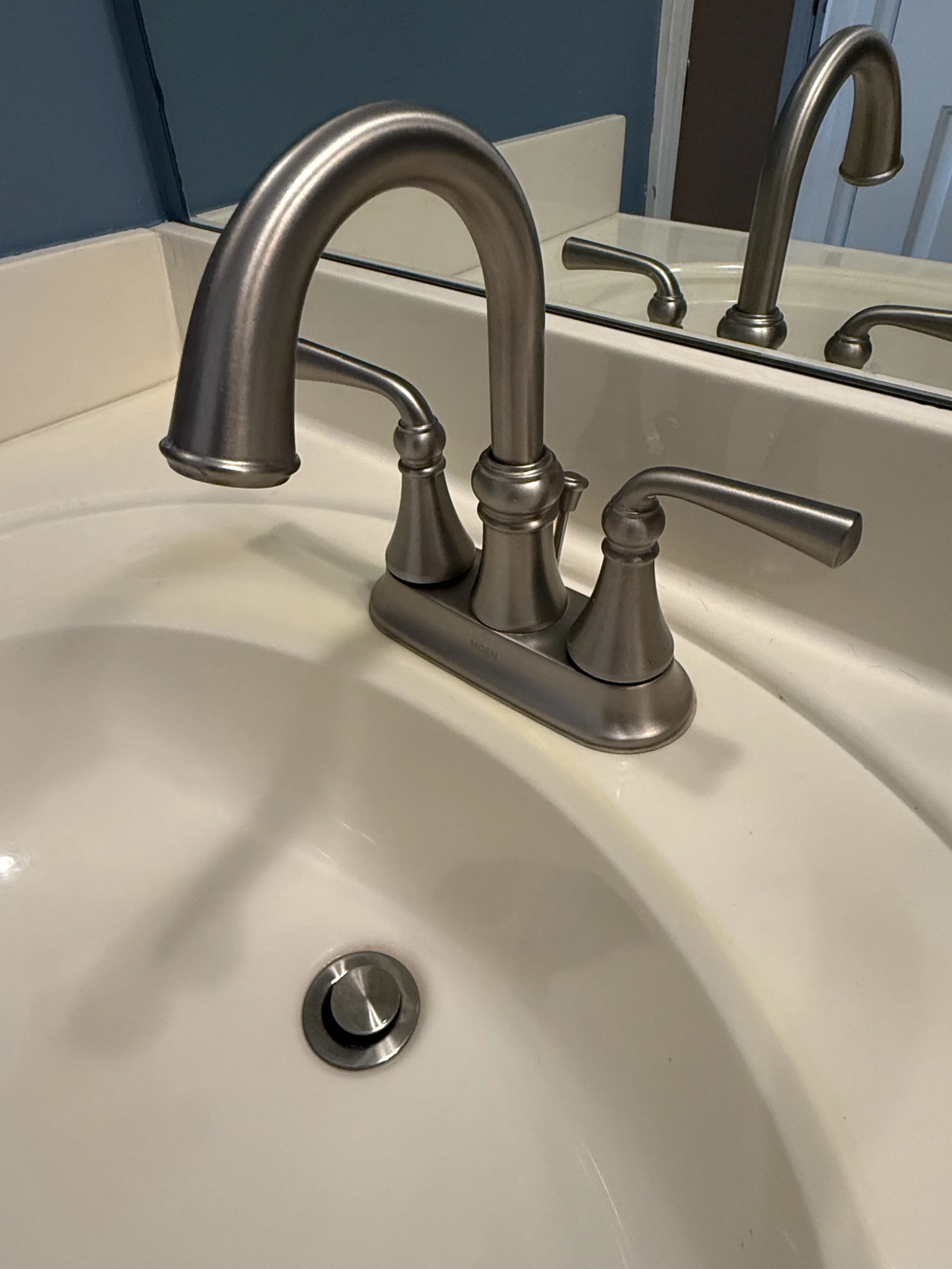 A bathroom sink with a brushed nickel faucet and a mirror behind it. The sink is white with a round drain at the bottom.