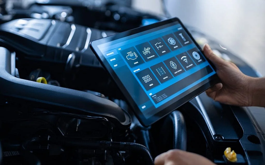 Person holding a tablet with a vehicle diagnostics interface, inside a car engine bay.